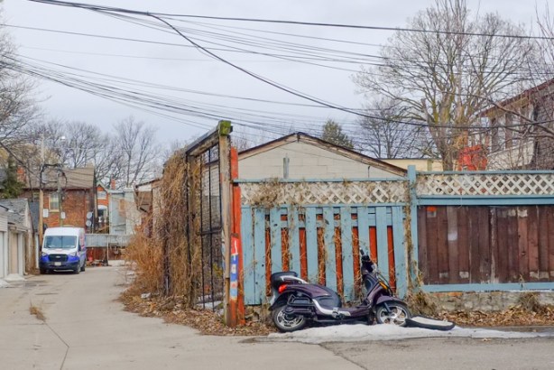 motorcycle parked in front of blue fence, beside a garage in a lane, near Queen Street West and Lansdowne