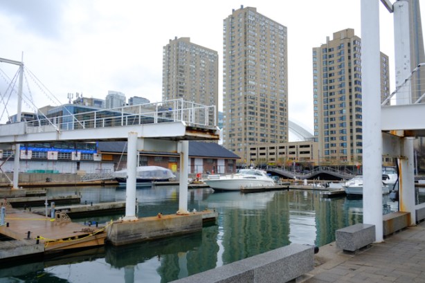Toronto highroses, condos near the water, marina and yacht club in the foreground with a couple of boats, pedestrian bridge over the water has been cut, impassable