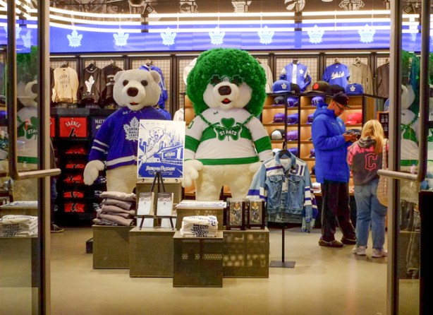 Looking in the door of the MLSE store at Scotiabank arena, and its Toronto Maple Leafs display, Carlton Bear in blue and white as well as in St. Pats white and green sweater, with green curly hair wig and green heart shaped glasses
