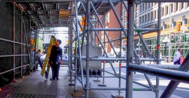 young man carrying a ladder, under scaffolding on sidewalk