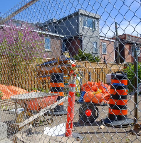 looking through chainlink fence to construction site with wheelbarrow as well as black and orange cones, orange plastic rolled up