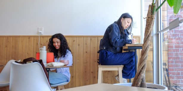 two women in a coffee shop, one is working at a table by the window, the other is seated at another table