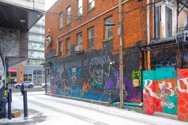 mural in an ossington alley, black background, text that says Designers of Canada, with three portraits, on the lower level of a three storey red brick building