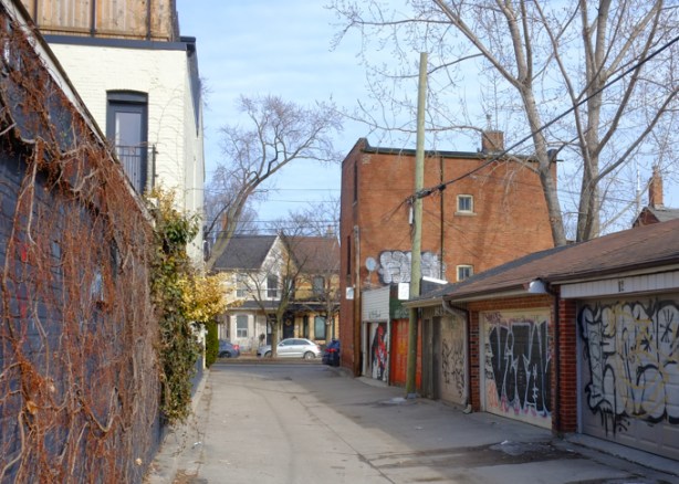 looking down an alley, garages with graffiti on the right, fence on the left, at the end, a semi divided house that is different on either side, also at end of alley, on right, a larger red brick building (old)