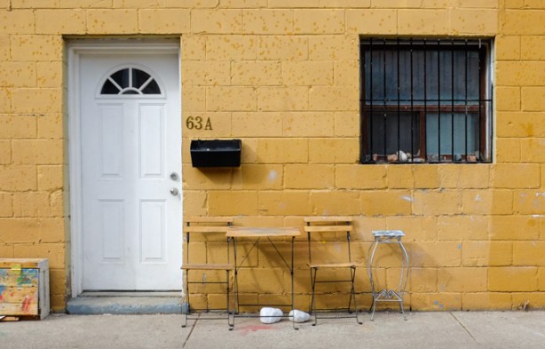exterior of building, concrete block painted a dark yellow,, white door, and black window, with 2 small chairs and a table, and a stool, immediately outside the building