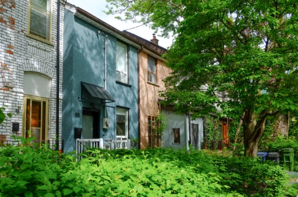 row houses, brick painted white house on left, then a blue house, and then brown, with a lot of green plants in the front yard. 