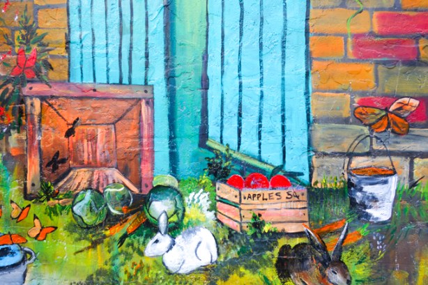 a white rabbit nibbles on the grass beside a wood crate full of red apples, part of a mural of nature scenes by B C Johnson
