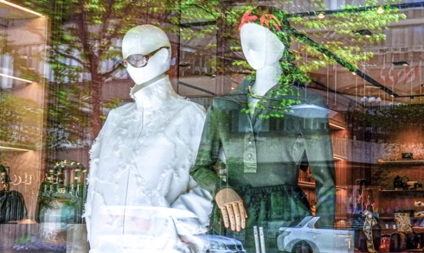 2 white mannequins in a window, one in dark glasses and the other in tight fitting cap, looking out a window, with reflections of trees and traffic