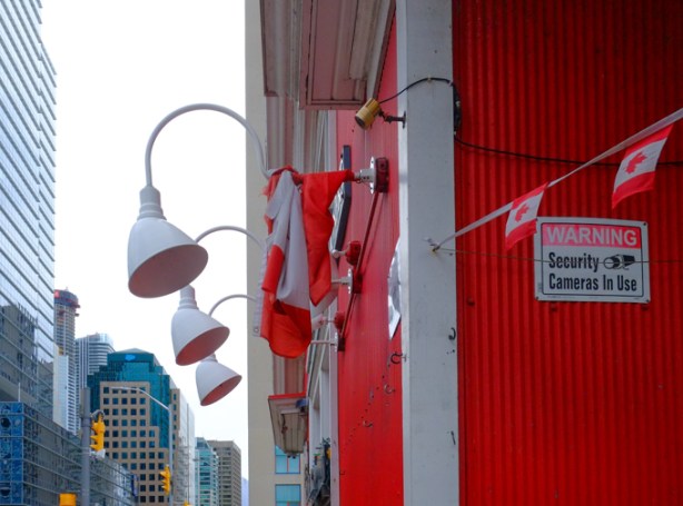 white lamps overhead on a red building, bedraggled Canadian flag on one of the lamps, a sting of smaller Canadian flags beside