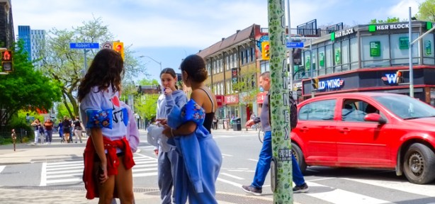 a group of people standing on the corner or Robert and Bloor as a red car turns right beside them