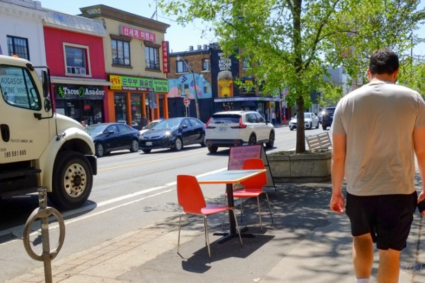 on the sidewalk, two small red plastic chairs wth a table between them, a man walking past, street scene in the background, Bloor Street