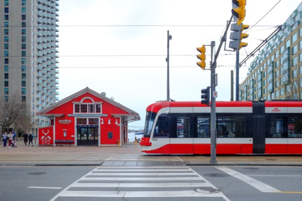 a red and white TTC streetcar on Queens Quay, traveling east past small red building that sells tourist stuff