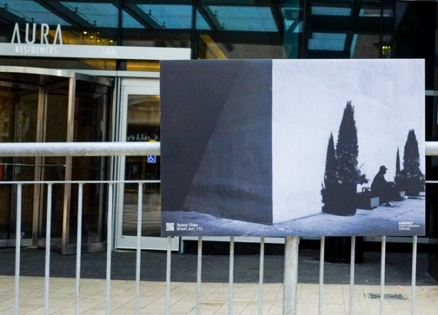 photograph by sunny chan mounted on metal fence in front of entrance to aura condo, image is of a man sitting on a bench beside a small pine tree, shadowy