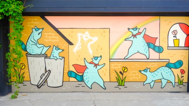 mural by Emily May Rose on a garage door in an alley, masked raccoons with red capes cavorting, one in a garbage can