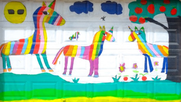 painting on a white garage door of rainbow striped unicorns under blue clouds and an apple tree