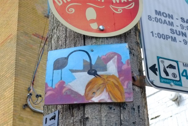 a little painting of a black bird with a long neck pecking at someone's brown hair, small painting mounted on a wood utility pole along with traffic signs, no parking