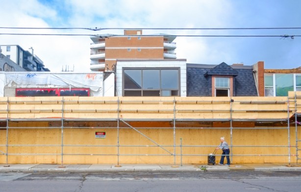 an older woman pushes her shopping under a passageway alonside plywood construction hoardings. Tops of old buildings about to be demolished can be seen on top