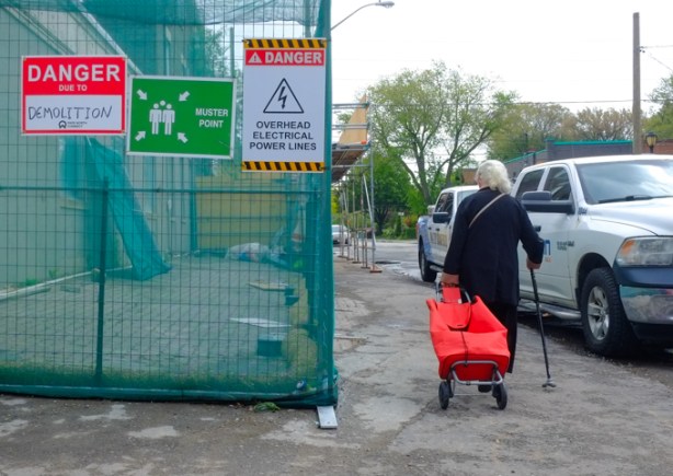 woman with a cane and pulling a shopping cart, walks past a construction site with green hoardings