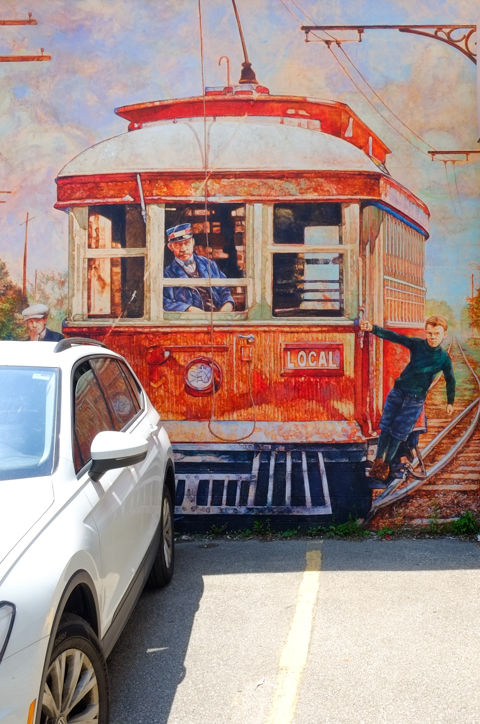 a white car is parked in front of one of the Islington heritage murals, boy hanging off side of a local train