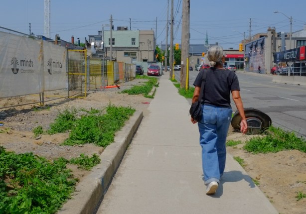 woman with gray hair, wearing jeans, walking on sidewalk part vacant lot with fence,