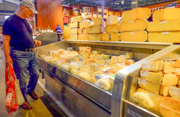 a man in jeans and blue shirt, holding red shopping bag, is looking at a display case full of different kids of cheese