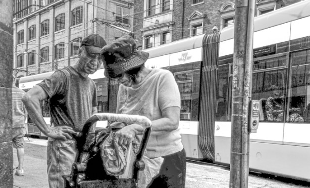 black and white photo of a couple standing on a street corner while a TTC streetcar passes them, both are wearing hats, she is looking for something in her bag and he is watching her