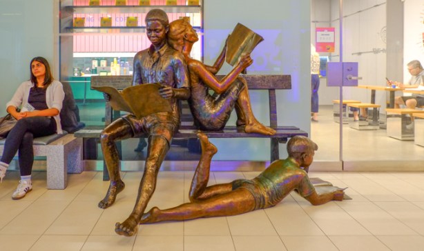 statue, sculpture, in post-media news building lobby, a group of 3 people reading, 2 sharing a bench, and one boy lying on the ground.