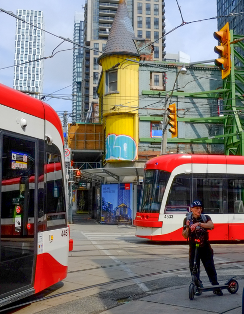 two streetcars going in opposite directions about to pass each other at corner of church and queen, a man on a scooter is also in the intersection