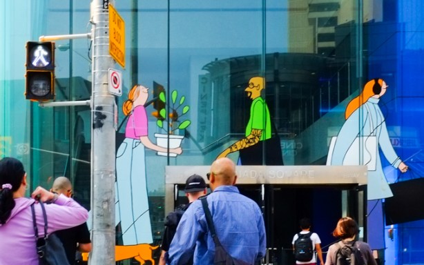 people crossing Eglinton Ave towards entrance to building on southwest corner of Yonge & Eglinton where there are images of people painted on the window