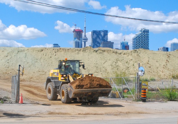 bulldozer, or front end loader with load of brown soil, heads out gate of construction site towards road, CN TOwer and toronto skyline in the distance
