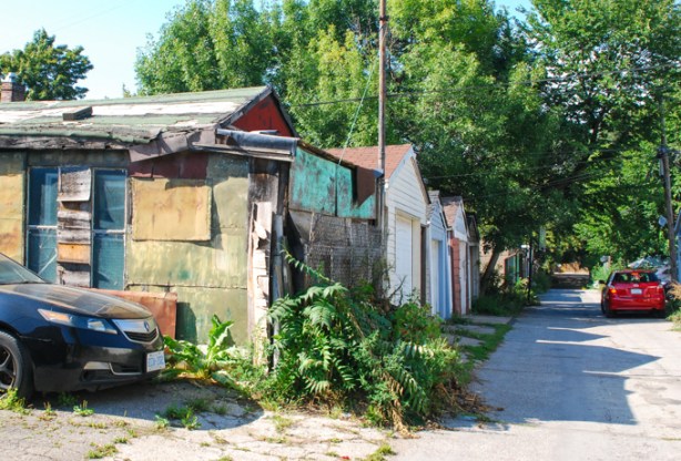 small red car parked in a lane, line of garages, tall trees,