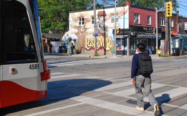 young man crossing Queen Street near Leslieville mural, and beside a TTC streetcar
