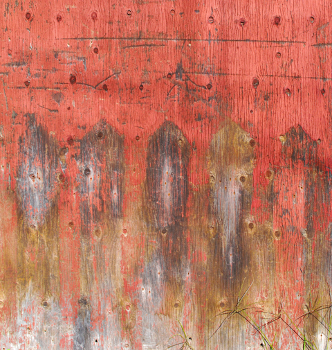 old plywood on a fence, once painted red, now faded to slightly orange red, with dirt splotches and worn areas, texture, pattern, abstract
