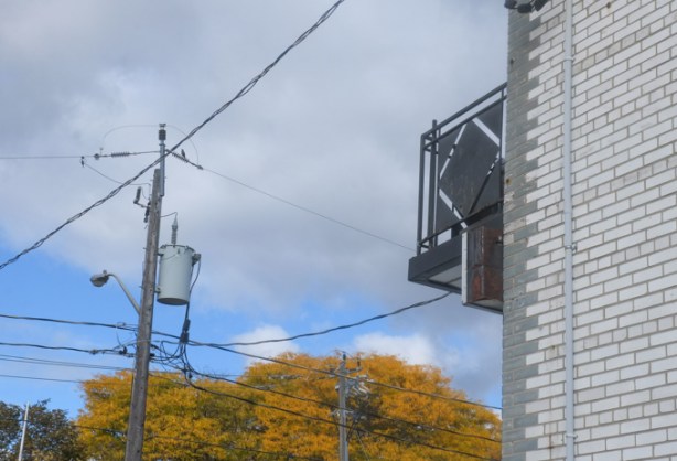 balcony wth black railing on a grey brick building, blue sky with a cloud, transformer on hydro pole, with some wires