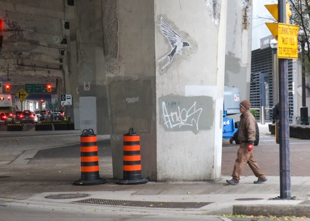 man in brown jacket is about to walk under the gardiner expressway, past a concrete pillar with a black and white picture of a bird in flight, street art, 