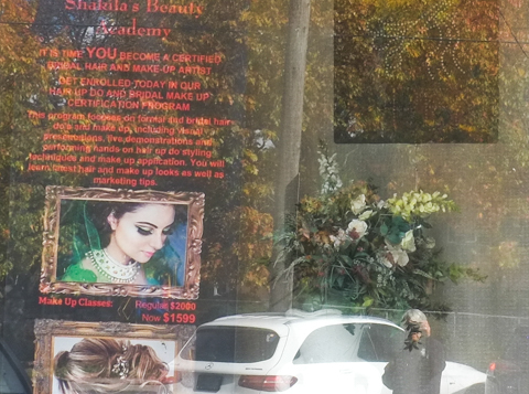 looking in the window of Shakira Beauty Academy, with some reflections of trees and cars outside