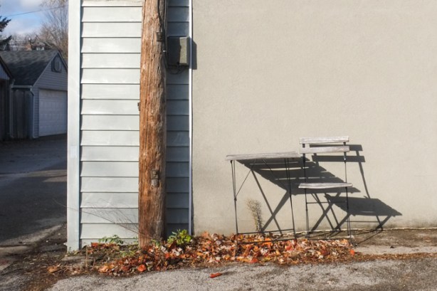 small table and chair outside, agains beige wall, shadows, autumn leaves on ground, sunny day, alley view