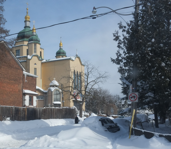 church, on snowy street, with cars parked but under snow cover, a woman walks past
