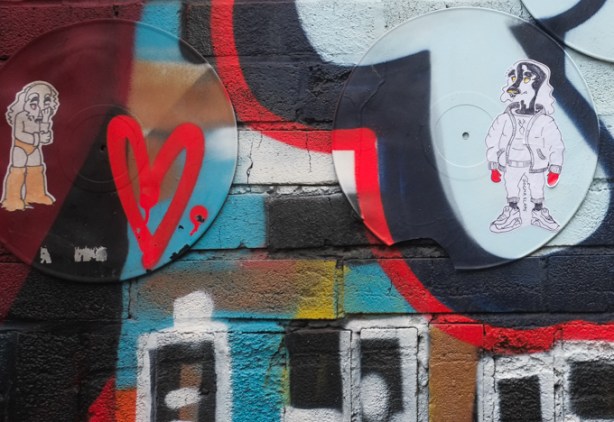 2 slaps graffiti on a wall already covered with street art paint. One is a dog, standing on two feet and dressed in human clothes - or perhaps it is a man with a dogs head