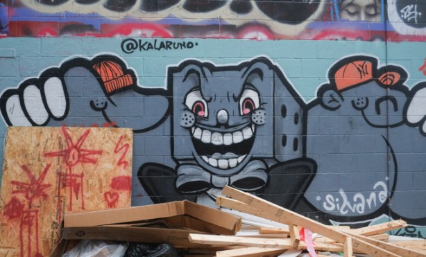 lumber and trash in front of a mural in graffiti alley, grey squarish character with human face by Kalaruno