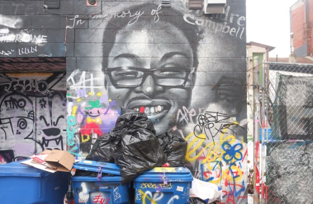 blue trash bins and black garbage bags piled in front of street art mural, black and white portrait of a man with short dark hair and glasses, with text that says in memory of Andre Campbell
