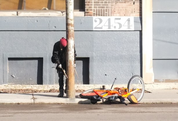man leaning against a metal utility pole on sidewalk, a stick in each hand, bicycle lying on the ground beside, banging on pole with sticks