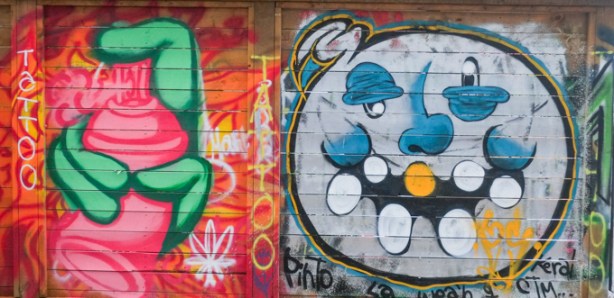 side by side, two smaller street art painting, graffiti alley, on left is green hand and pink spray paint can while on right is a stupid round face with big oval teeth