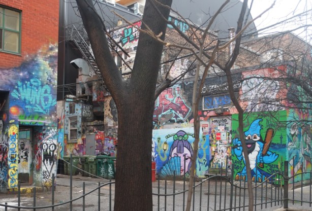 tree in front, looking across alley to side of building, completely covered in street art painting, graffiti alley