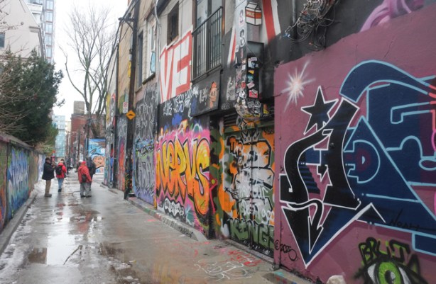people walking down a wet alley, puddles and reflections on the pavement, of the street art and graffiti on the surrounding walls