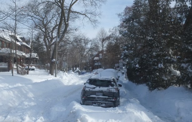 car parked on snow covered street, evergreens, as well as large bare trees, some houses,
