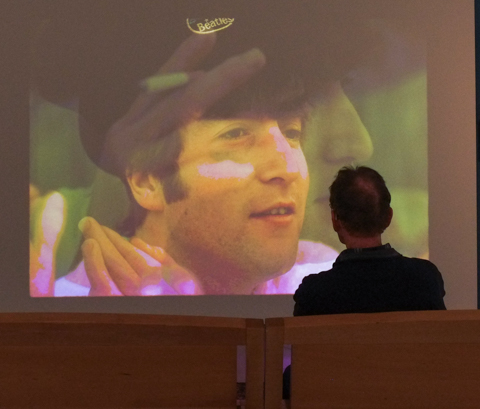 man with back to camera, sitting, watching a slide show with large image of the Beatles, AGO