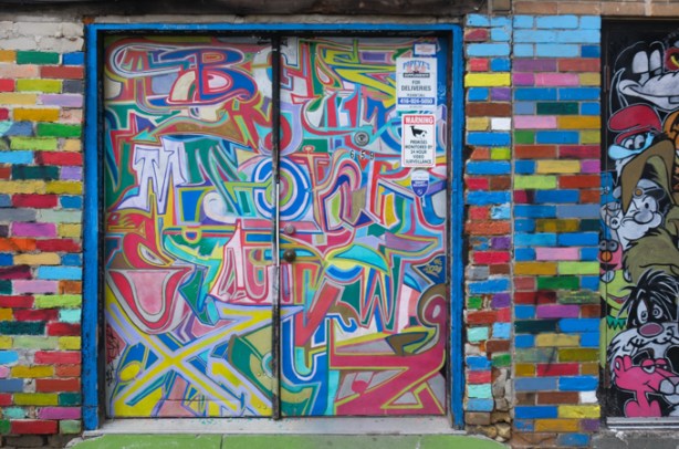 double door in alley, covered with street art which is letters of the alphabet all jumbled up together in many colours
