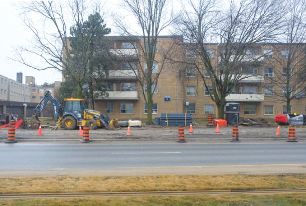 digger and construction equipment outside a 4 storey brick apartment building, rainy day, trees in front of building