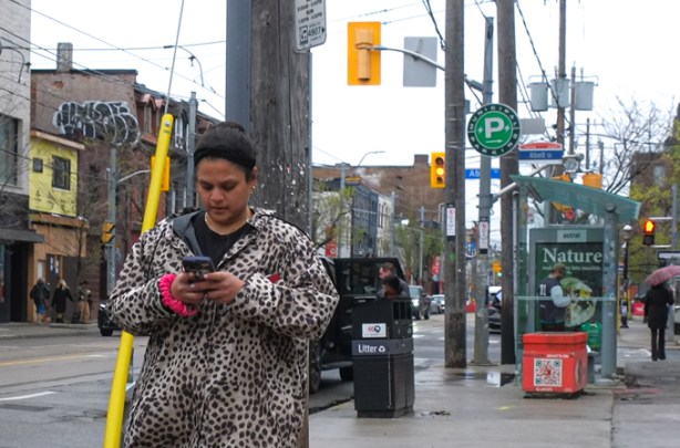 a woman with short black hair, wearing a long leopard print jacket, walks along queen street west while looking at her phone